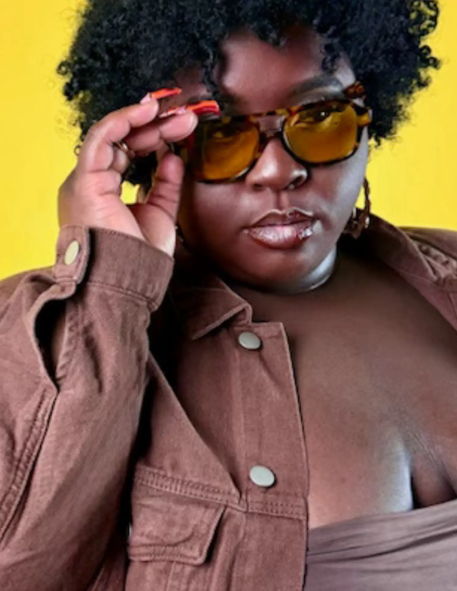 Close portrait of a woman peering over amber glasses against a bright yellow backdrop